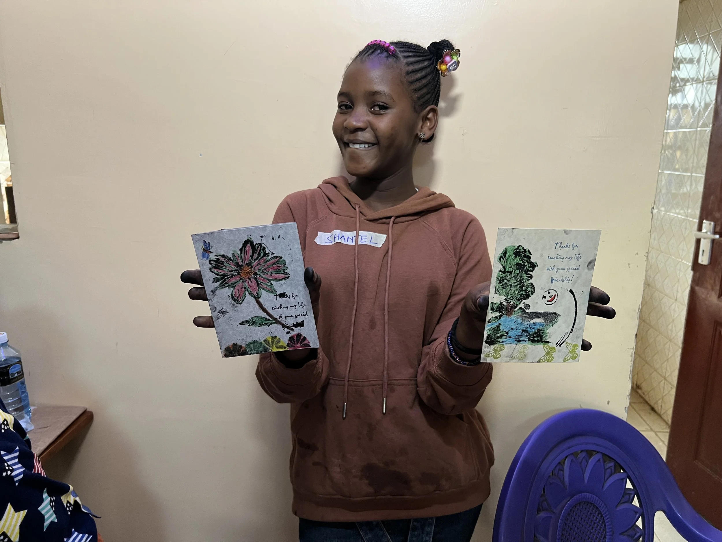 A young girl with braided hair decorated with colorful beads, smiling and holding two handmade greeting cards indoors against a beige wall.