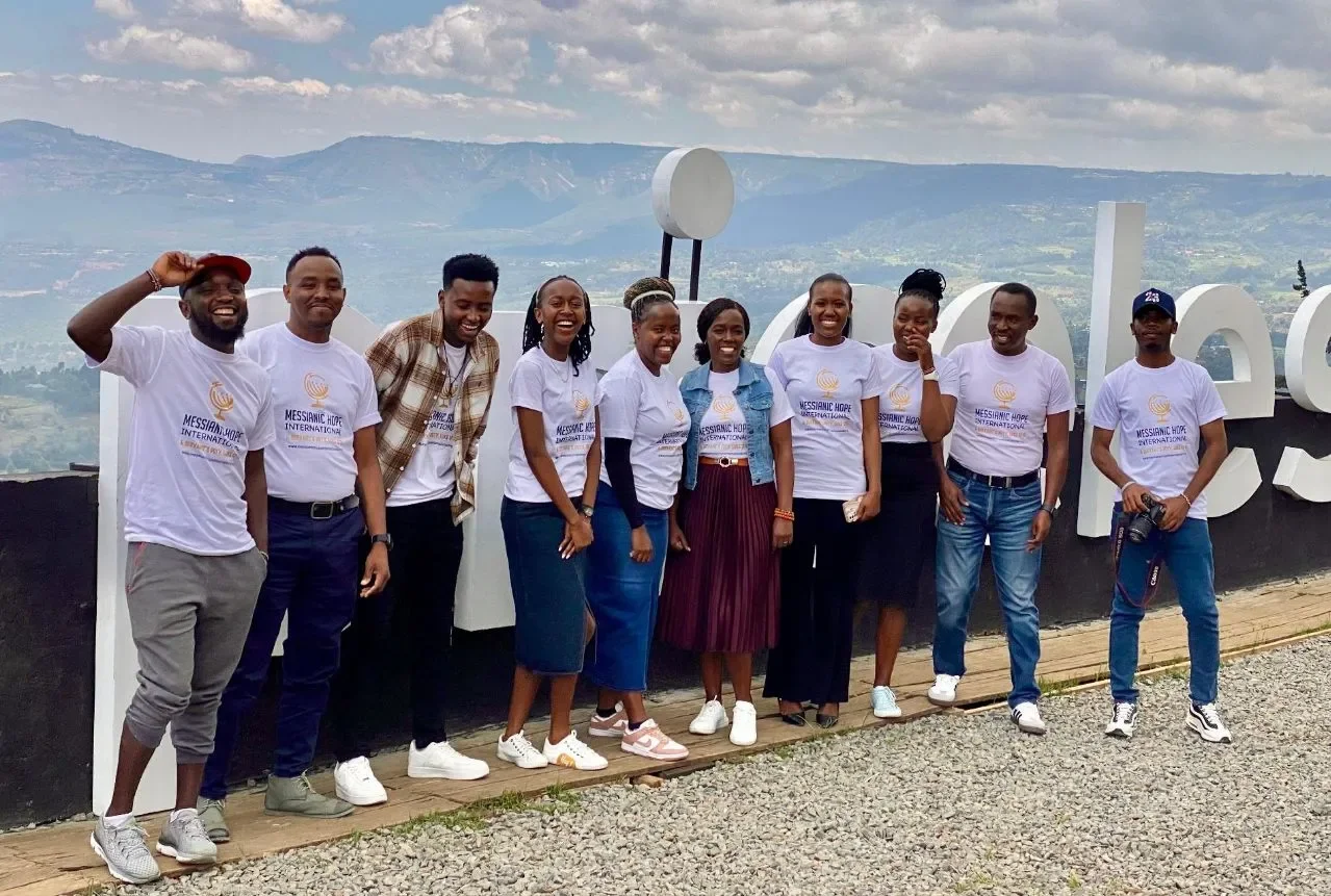 Group of ten people standing outdoors in front of a scenic view and large white letters spelling out 'Hope.' Some are wearing white T-shirts with a logo and text that reads 'Messianic Hope International.' The group is smiling and dressed casually.