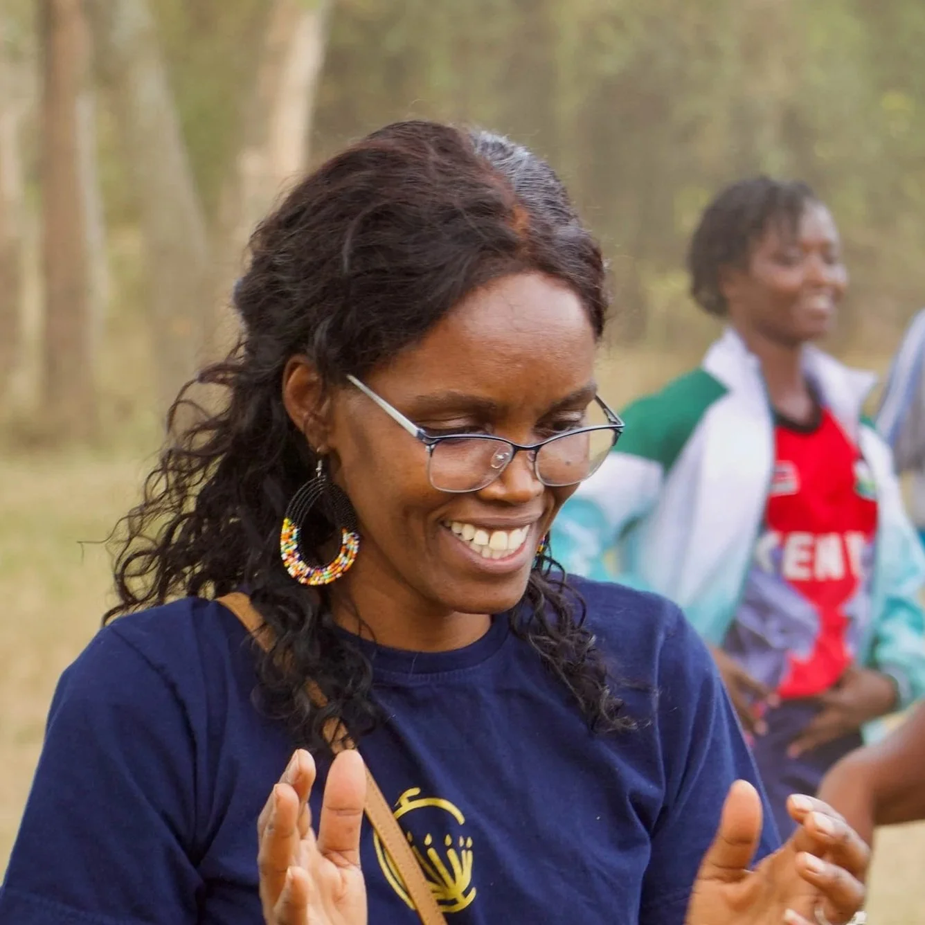 Smiling woman with glasses and large earrings enjoying an outdoor event with others in the background.
