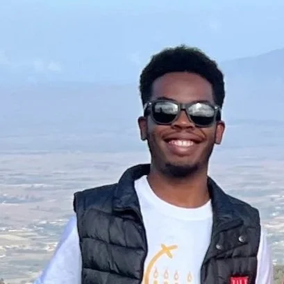 A young man smiling outdoors, wearing sunglasses, a black puffer vest, and a white t-shirt, with a mountainous landscape in the background.