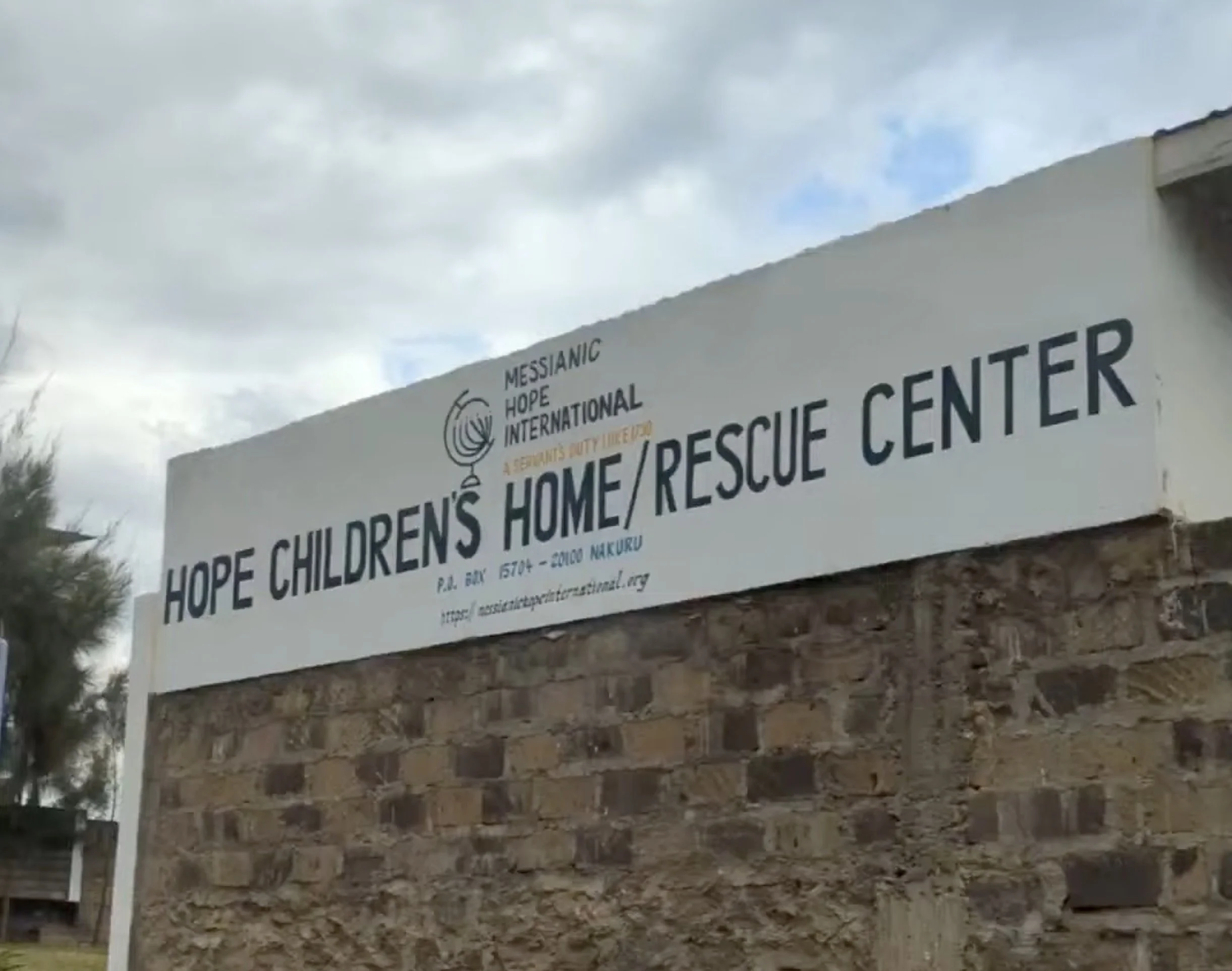 Sign for Hope Children's Home/Rescue Center with Messianic Hope International logo, on a brick wall under cloudy sky.