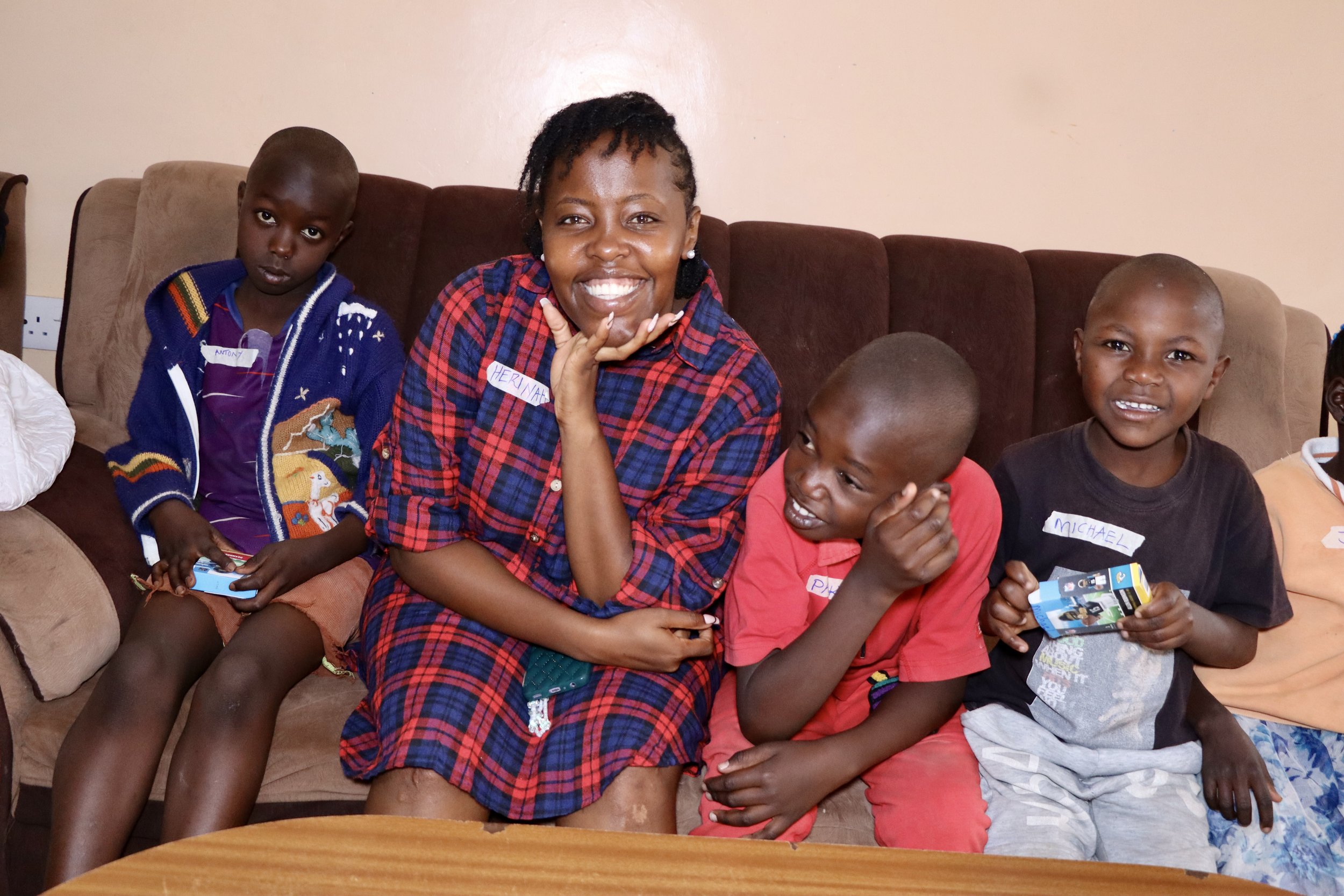 A group of children and a woman sitting on a brown couch indoors, smiling and holding small items, with some wearing name tags.