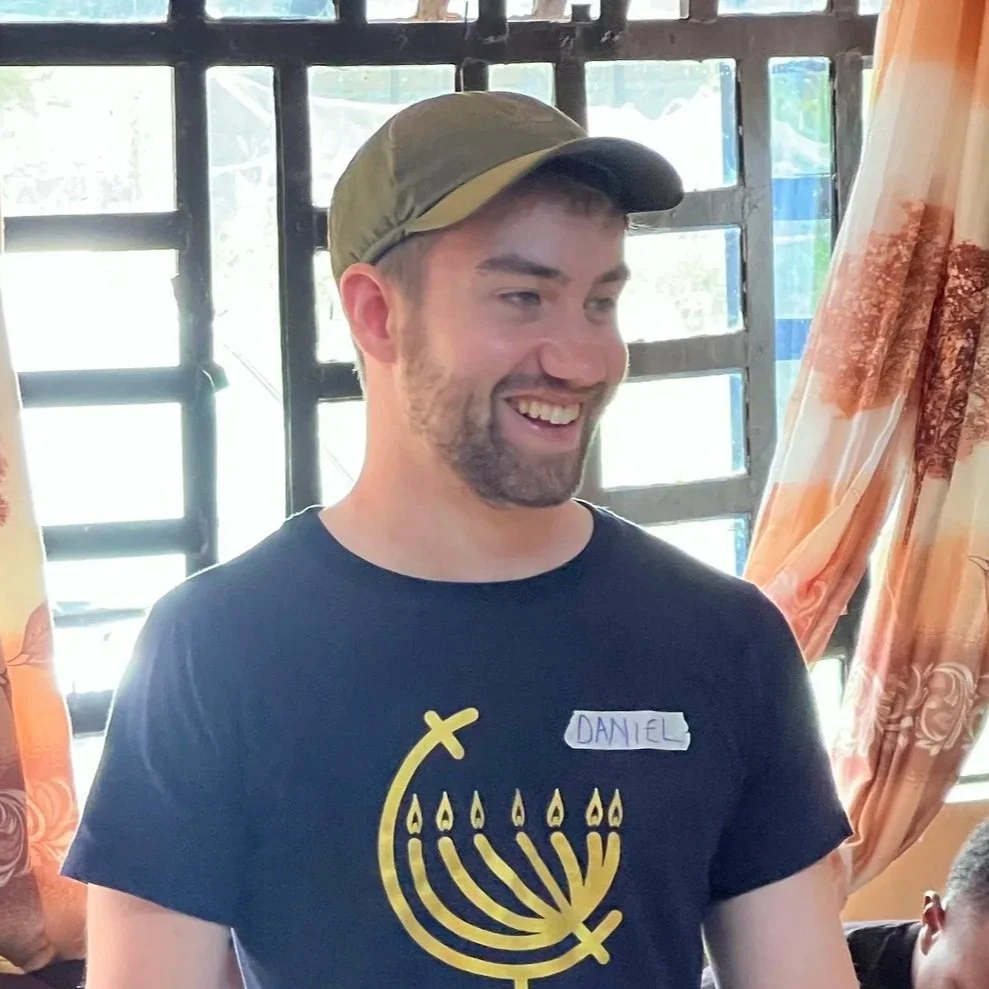A young man with a beard, wearing a green cap and a black T-shirt with a yellow menorah design, smiling indoors near a window and curtains.