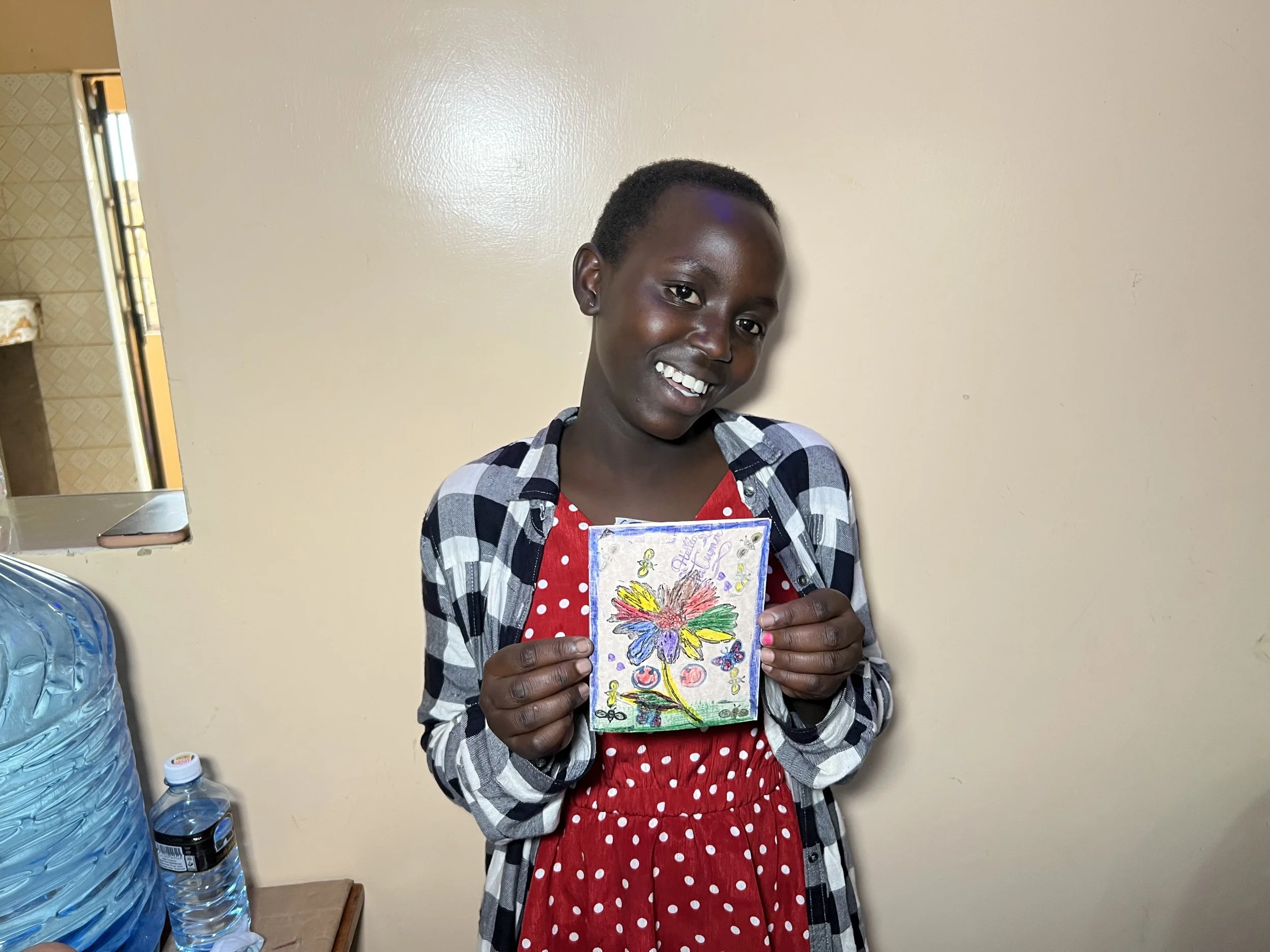 A smiling girl with short hair holding up a colorful drawing of a flower. She is wearing a red polka dot dress and a black, white, and gray checkered jacket, standing in front of a beige wall.