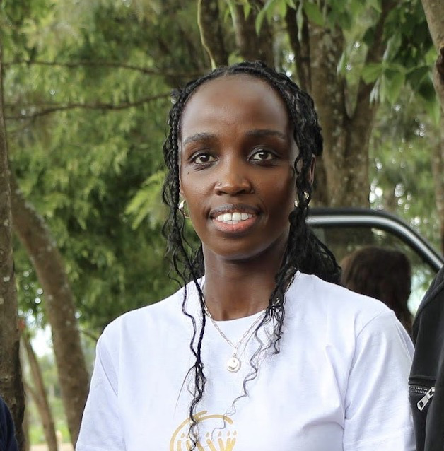 A woman with long black braided hair wearing a white t-shirt, standing outdoors with green trees in the background.