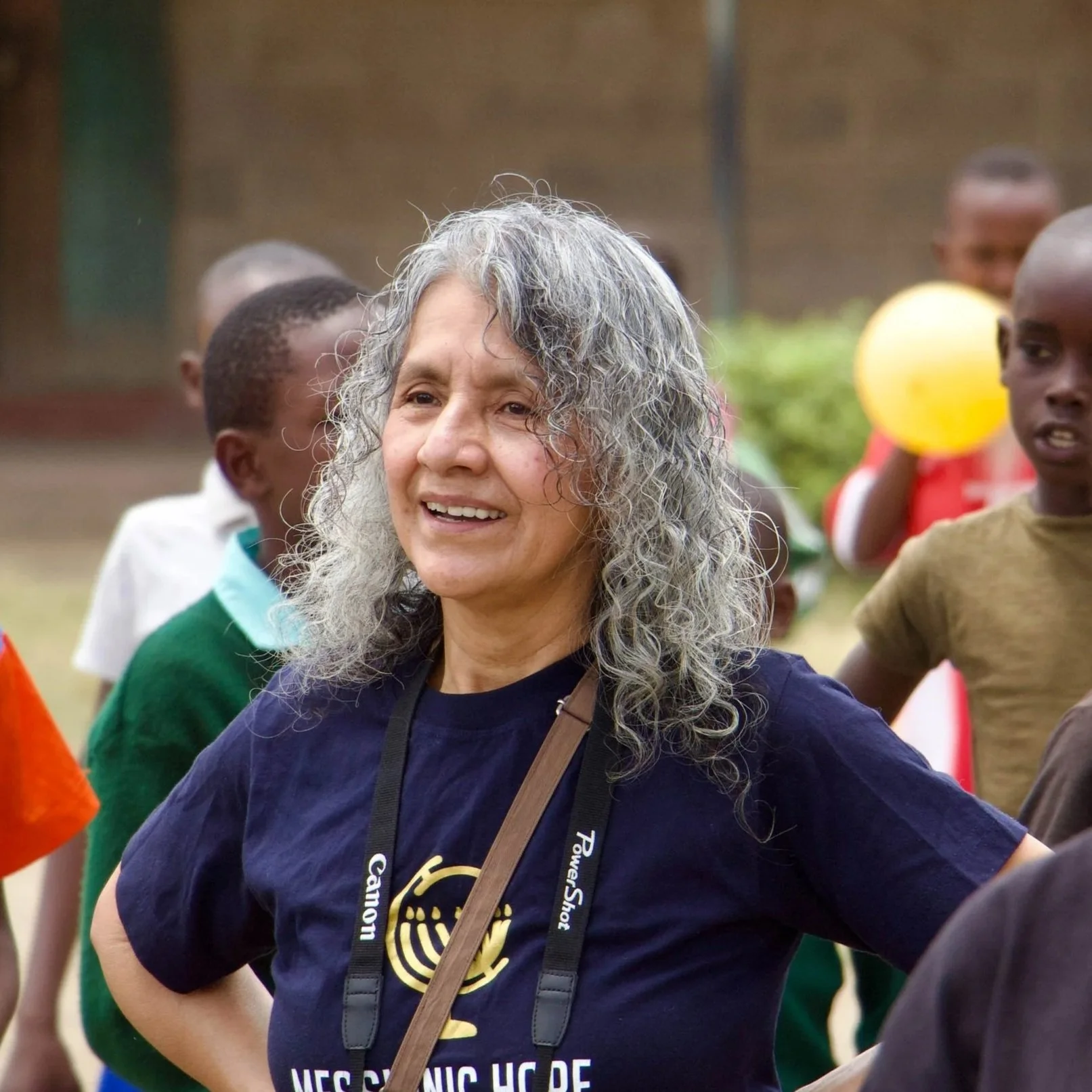 A woman with long, curly gray hair smiling at an outdoor gathering, surrounded by children, some holding balloons.