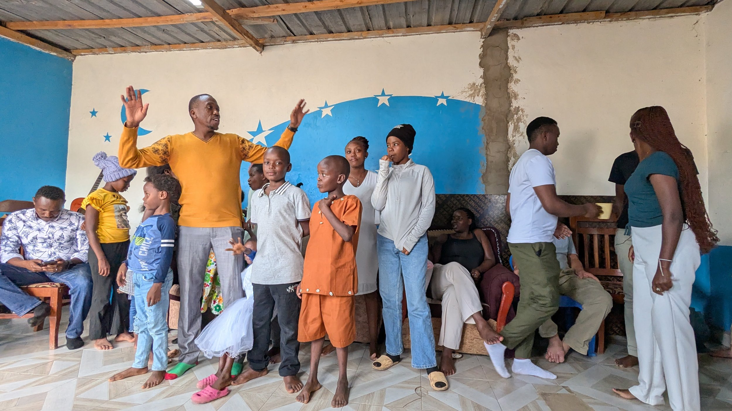 Group of children and adults gathered in a room with blue wall decorations, some standing and some sitting, engaging in conversation.
