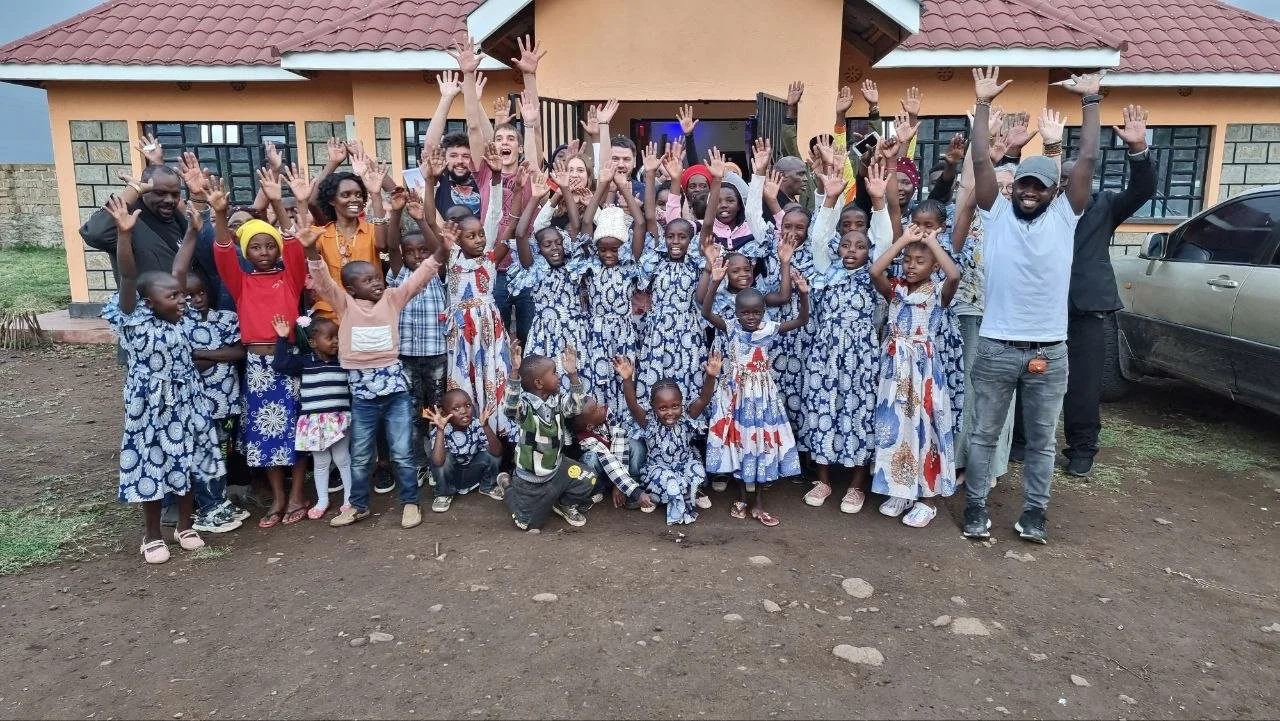 Group of children and adults standing outside a building, raising their hands and smiling.