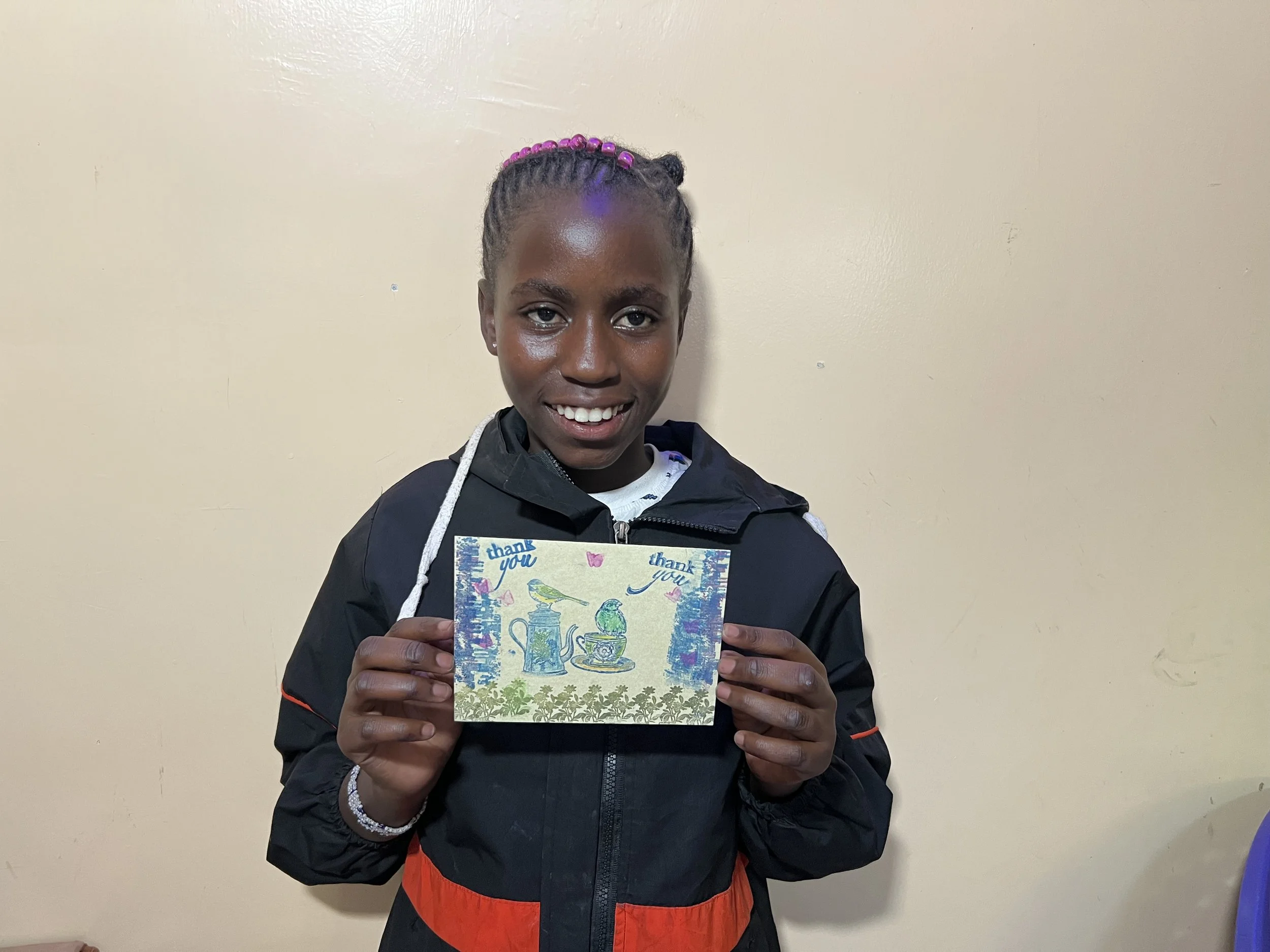 A young girl with braided hair holding a thank you card, smiling, standing against a beige wall.