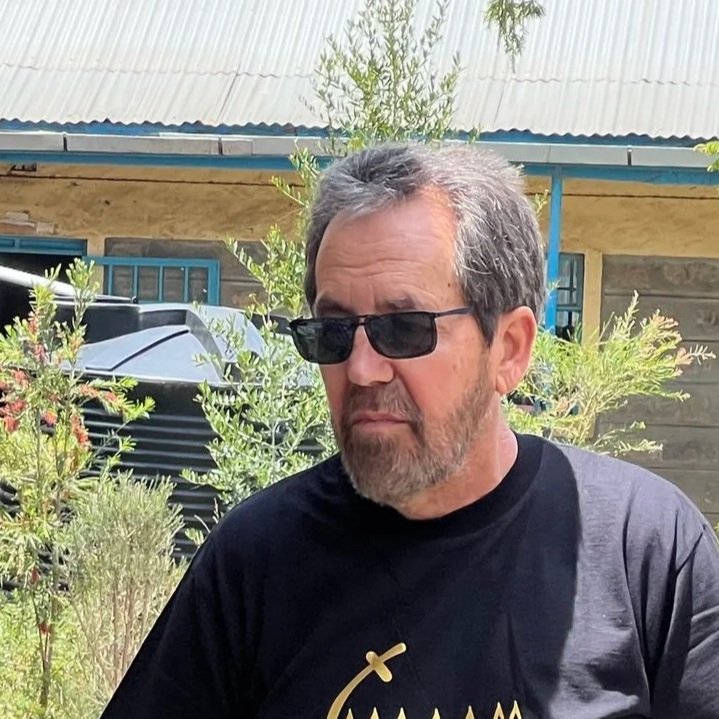A middle-aged man with sunglasses and a beard, wearing a black T-shirt, standing outdoors near greenery and a vehicle, with a building in the background.