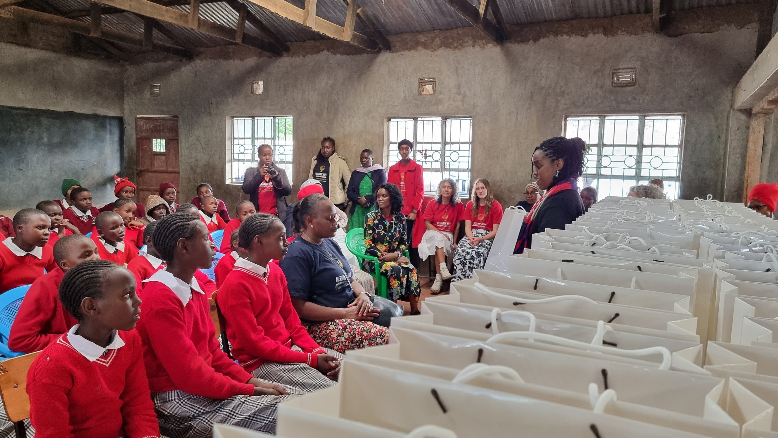 Group of children and adults in a classroom setting with a row of white bags in the foreground, some people wearing red, inside a rustic room with exposed wooden beams and barred windows.