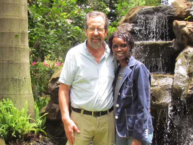 A man and woman standing together near a waterfall in a garden with greenery and rocks.