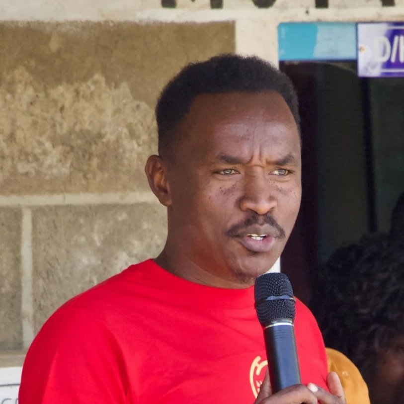 A man wearing a red shirt holding a black microphone and speaking outdoors.