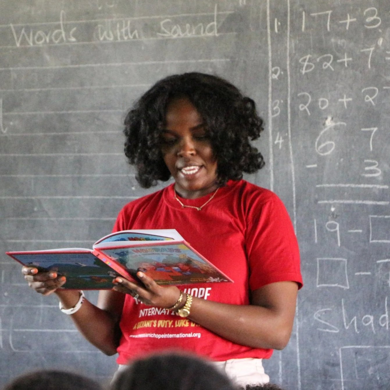 A woman with curly black hair wearing a red shirt reading a colorful children's book in a classroom with a chalkboard behind her.