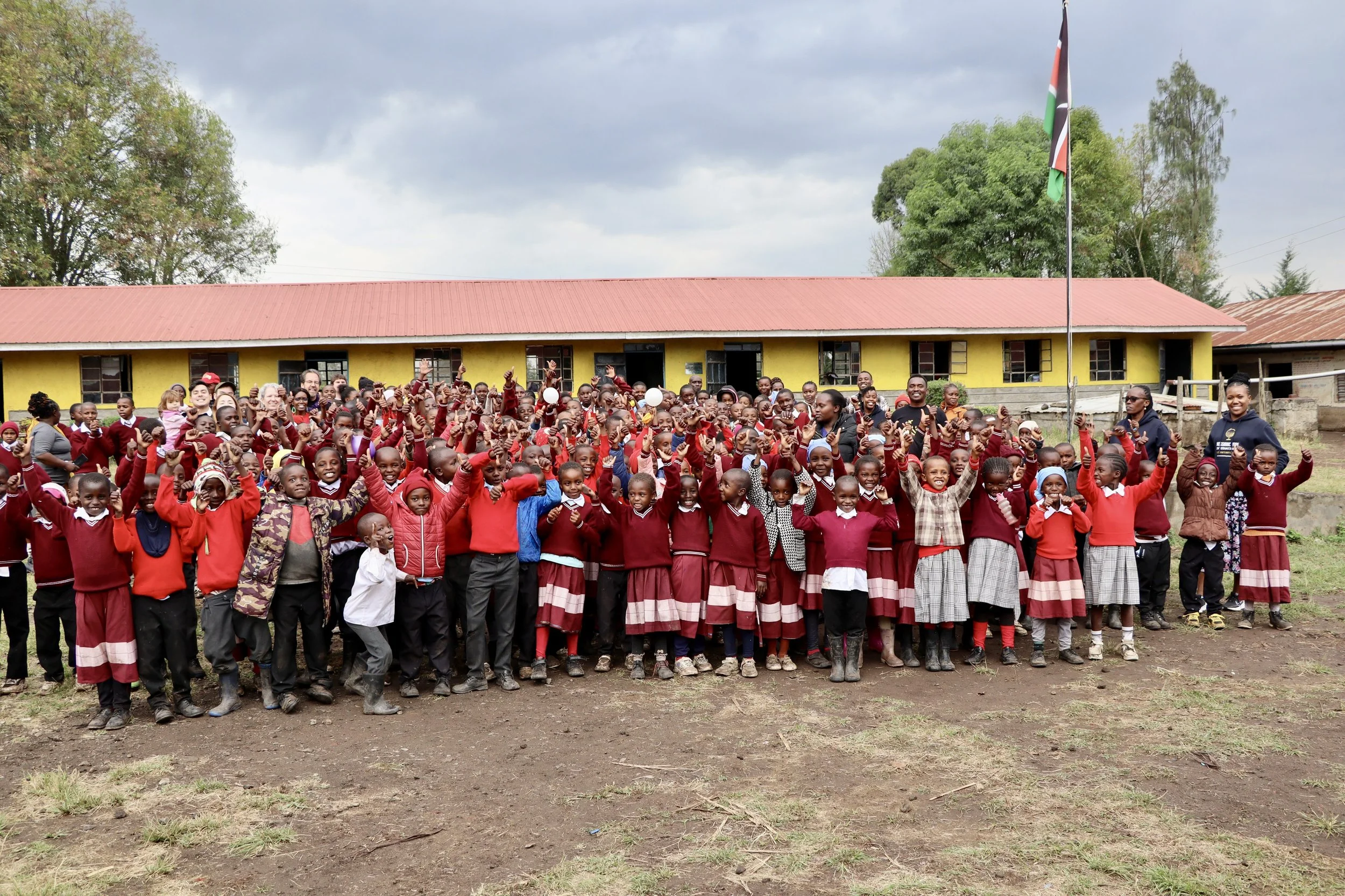 A large group of school children and teachers standing outdoors in front of a yellow school building, raising their hands and smiling. The children are dressed in red school uniforms, and there is a flagpole with the Kenyan flag nearby.