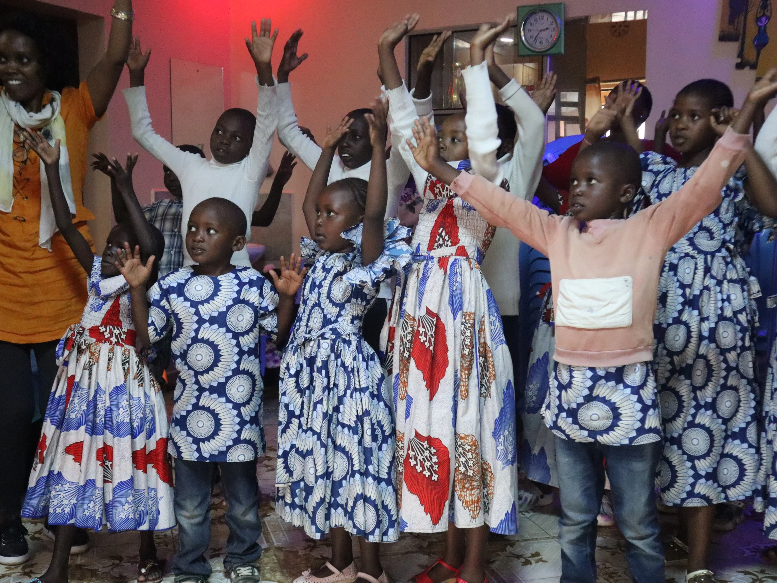 Group of children and a woman dancing and raising their hands indoors.