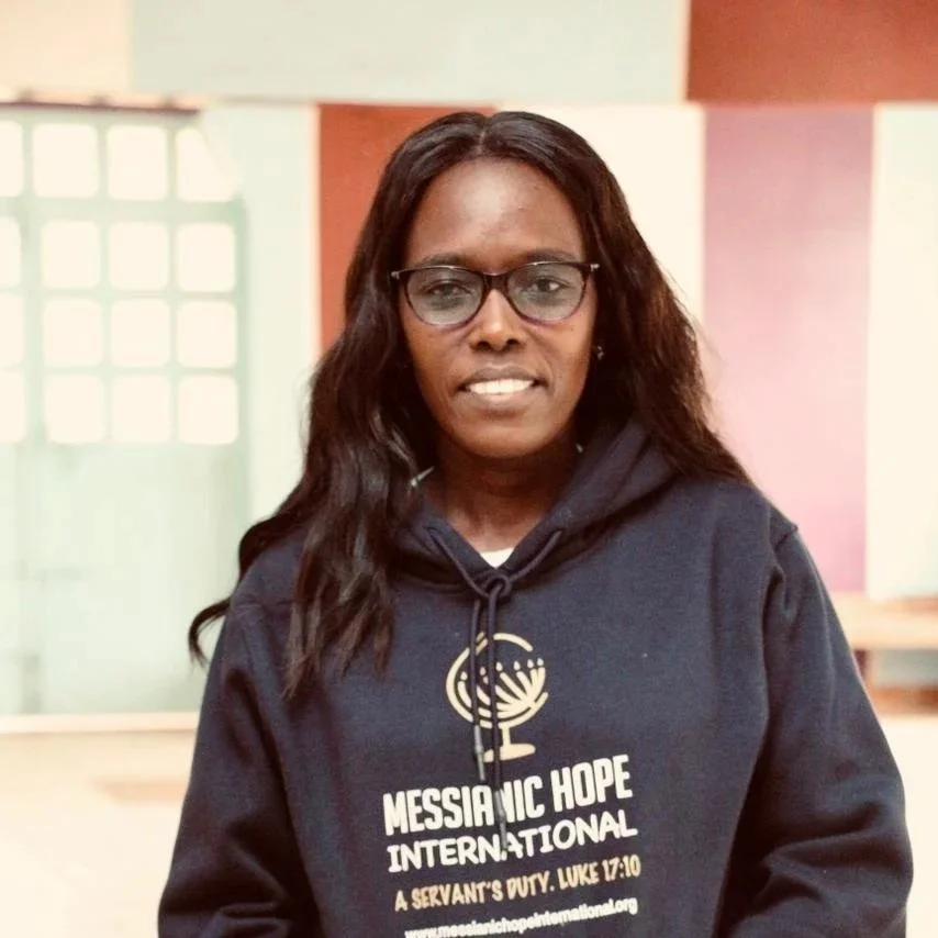 A woman wearing glasses and a navy blue hoodie with 'Messianic Hope International' logo and text, standing indoors with light green, white, and peach-colored walls in the background.