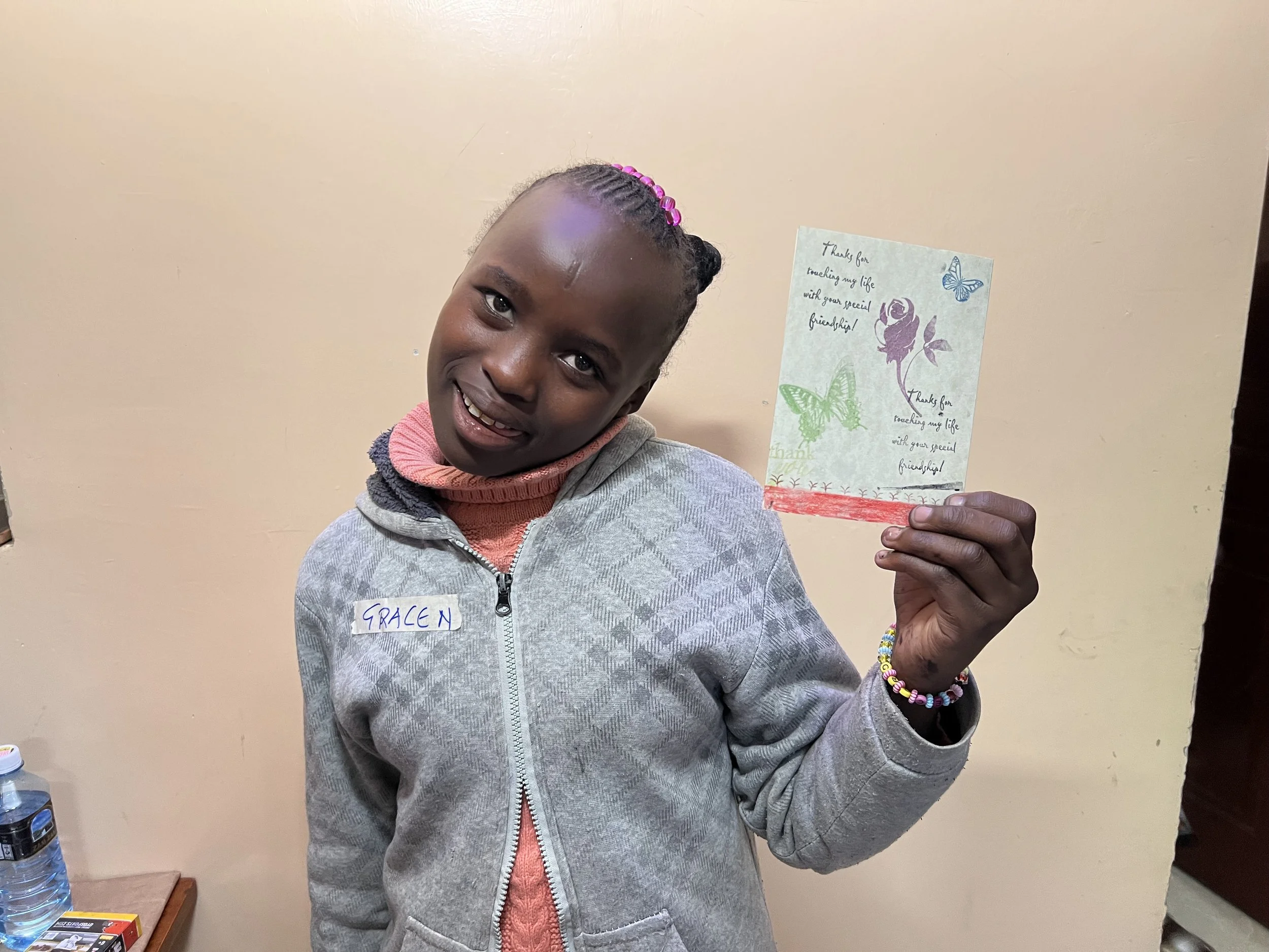 A young girl holds a handmade thank you card decorated with butterflies and a flower, smiling at the camera. She wears a gray jacket with a pink and peach sweater underneath, and has a colorful bracelet on her wrist. Her name tag reads 'GRACEN'.