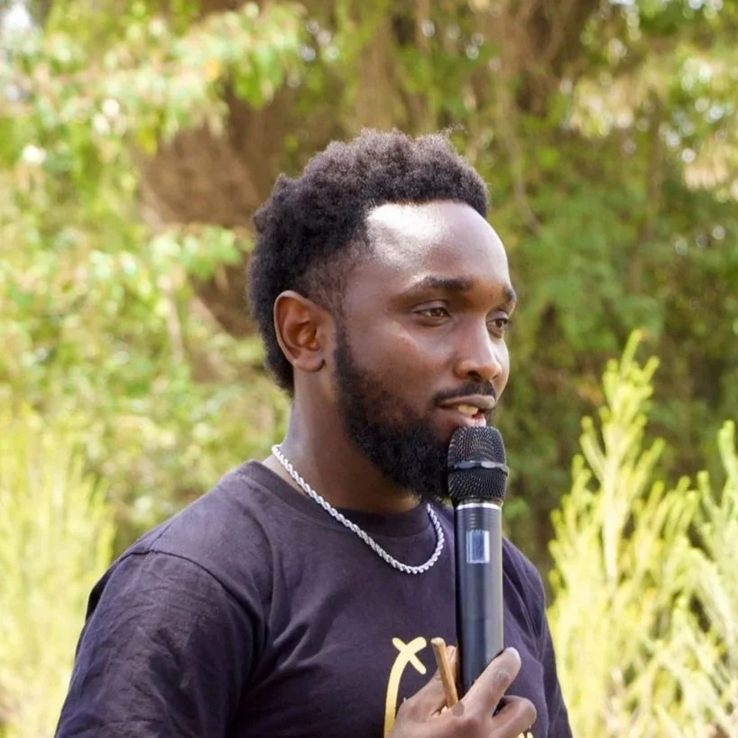 Young man with a beard and short curly hair holding a microphone outdoors, wearing a black T-shirt and a silver necklace, with green trees and foliage in the background.