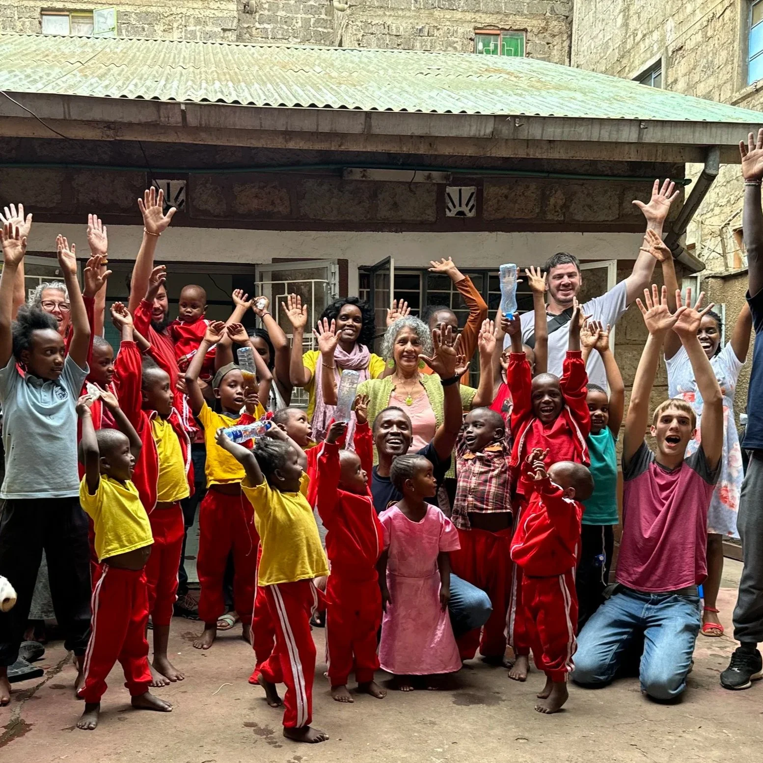 A diverse group of children and adults smiling and raising their hands in celebration outdoors in front of a building with open windows. Some children are wearing red and yellow outfits, and adults are dressed casually. The atmosphere appears joyful and lively.