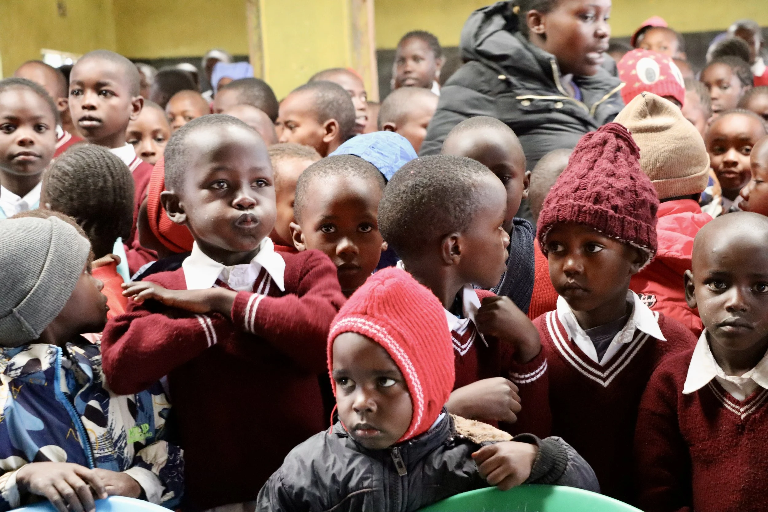 Group of children crowded together indoors, some wearing red sweaters, hats, and school uniforms, with expressions ranging from curiosity to seriousness.