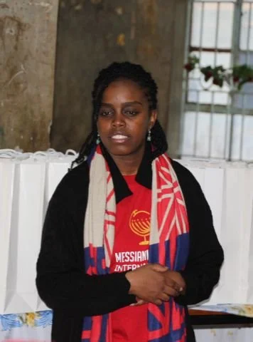 A woman with dark hair and dark skin wearing a red shirt with white text and a logo, along with a black sweater, standing indoors near a window and a plant.