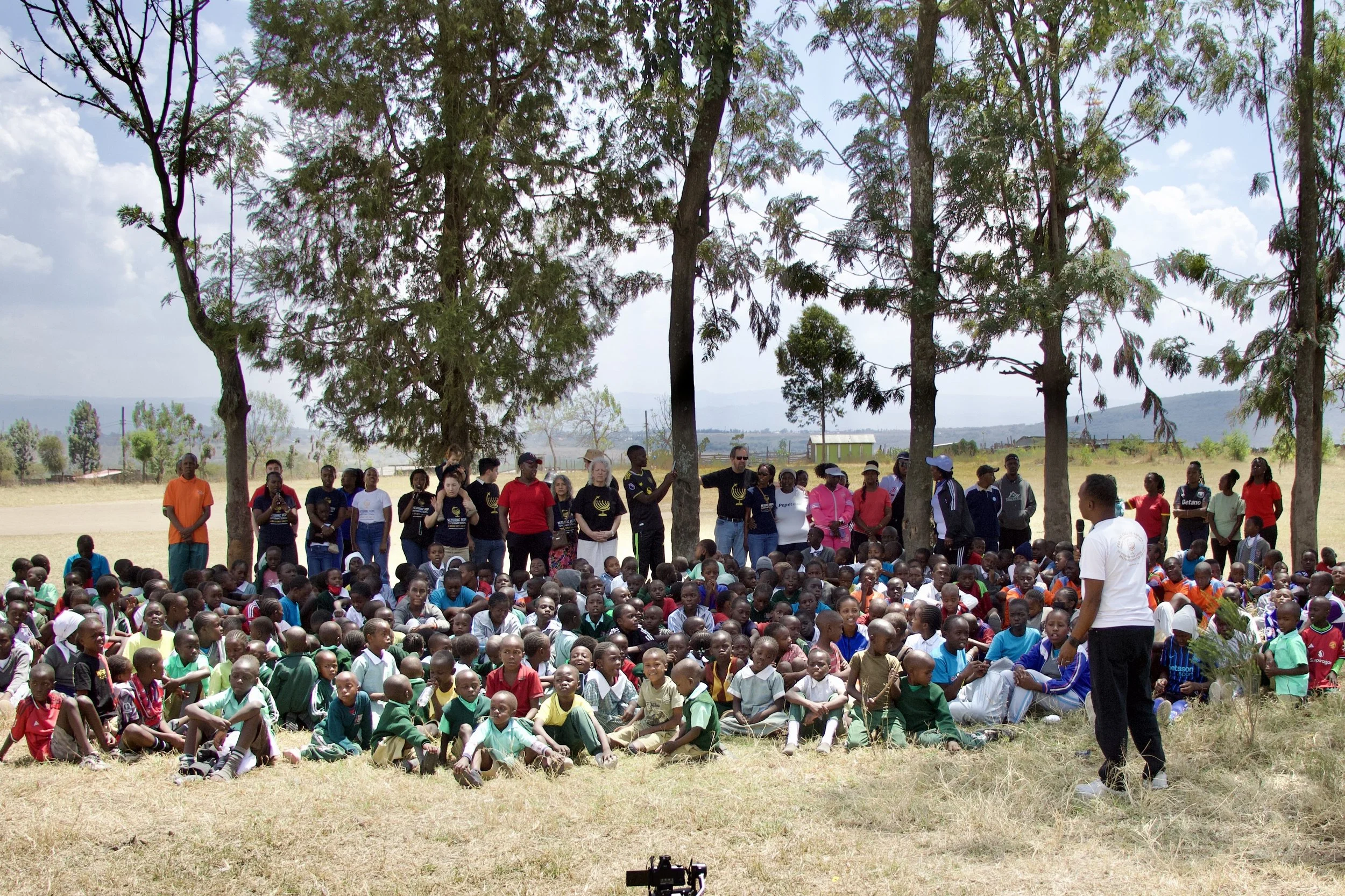 A large outdoor gathering of children and adults under trees, with a person standing in front speaking to the crowd, on a sunny day with open fields in the background.