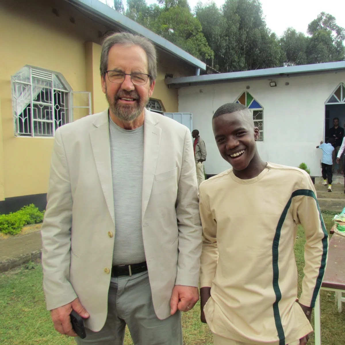 A man with glasses and a beard wearing a light-colored blazer and gray shirt standing next to a young boy in a beige sports jersey outdoors.