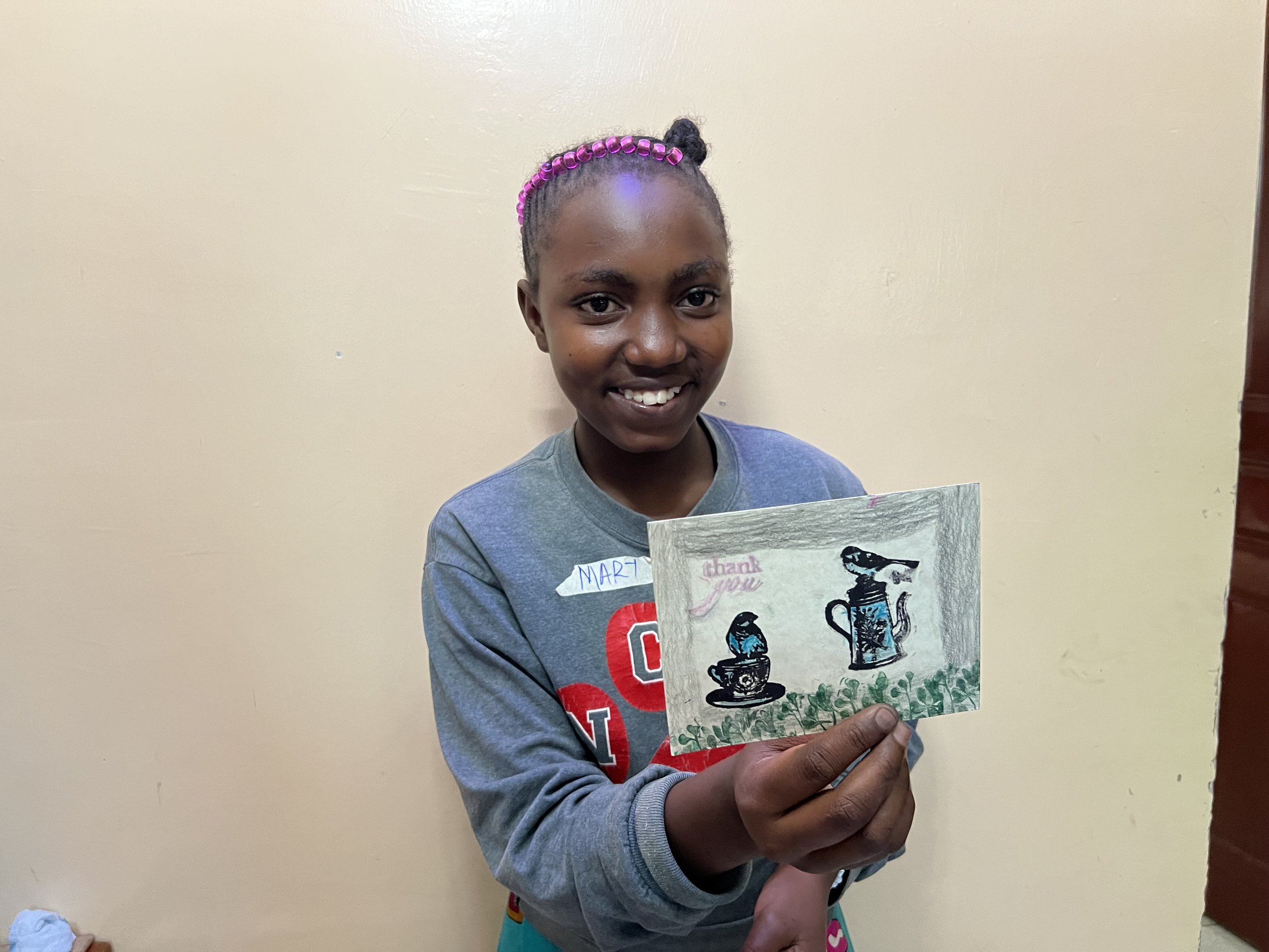 A young girl smiling and holding a handmade thank you card with drawings of teapots and cups.