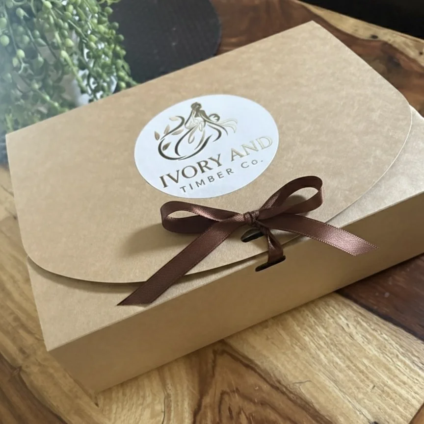 A beige cardboard box with a round logo sticker on the top that reads 'IVORY AND TIMBER CO.' with an illustration of a woman in a dress. The box is tied with a small brown satin ribbon. It is placed on a wooden surface with some green foliage in the background.