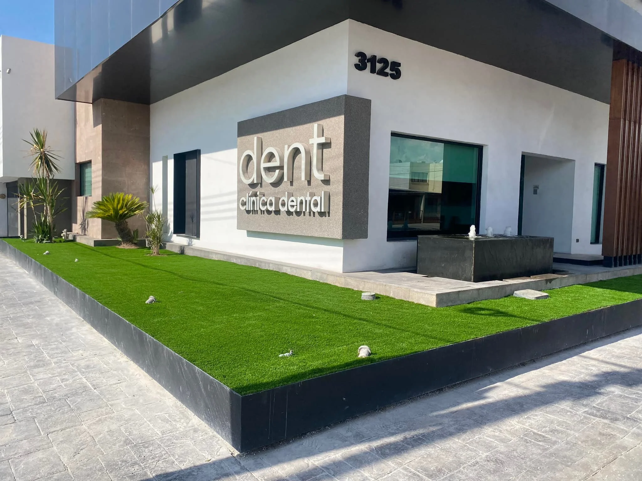 Fachada de una clínica dental moderna con señalización que dice 'dent clínica dental', ventanas grandes y un área de césped artificial con plantas decorativas