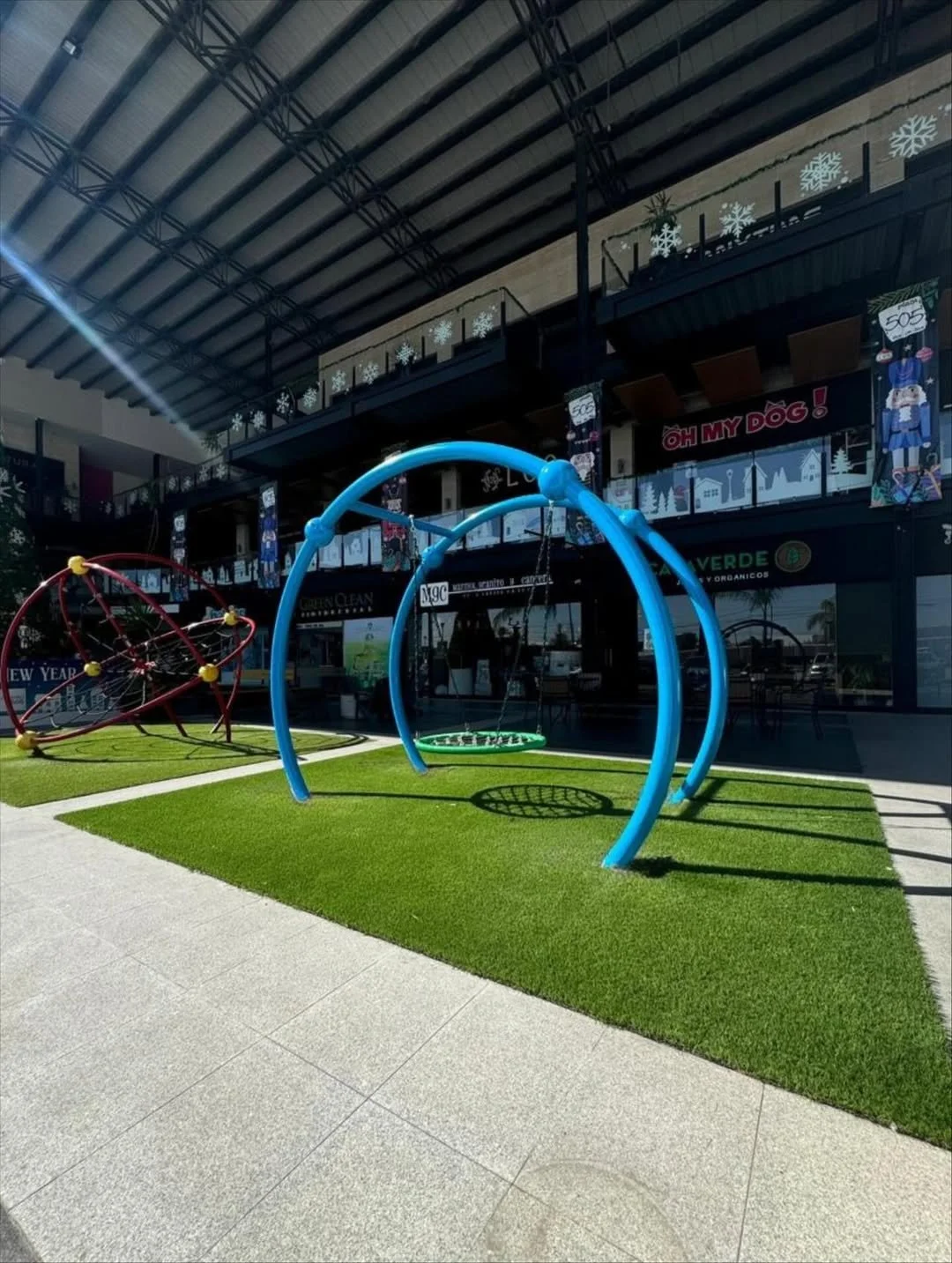 Área de juegos infantiles con columpios azules y una estructura de esfera metálica roja, en un espacio exterior con césped artificial y pavimento de baldosas, en un entorno comercial.