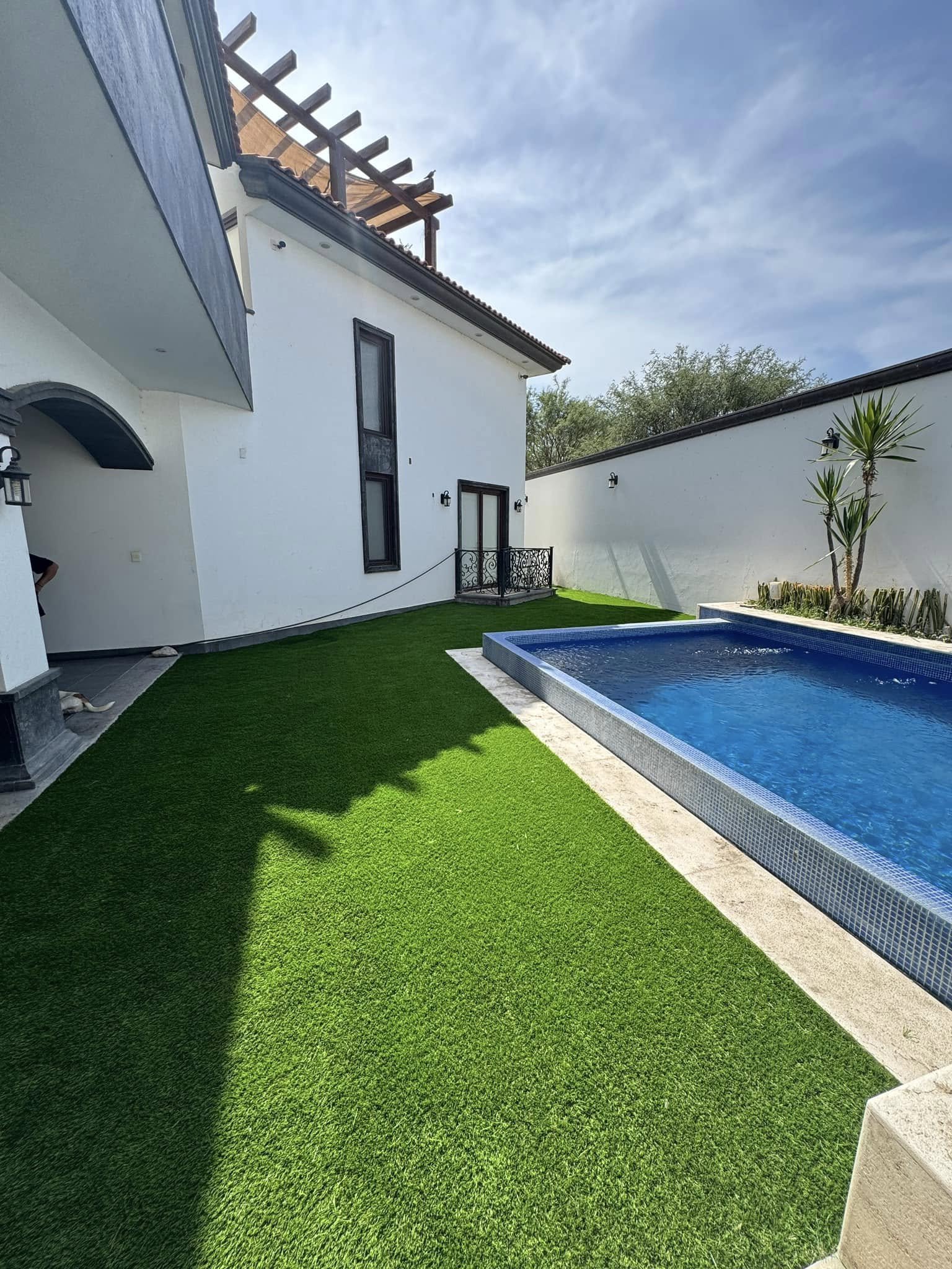 Jardín con césped artificial, piscina rectangular, muro blanco y una casa moderna de dos pisos con ventanas oscuras y un balcón de hierro black en la casa.