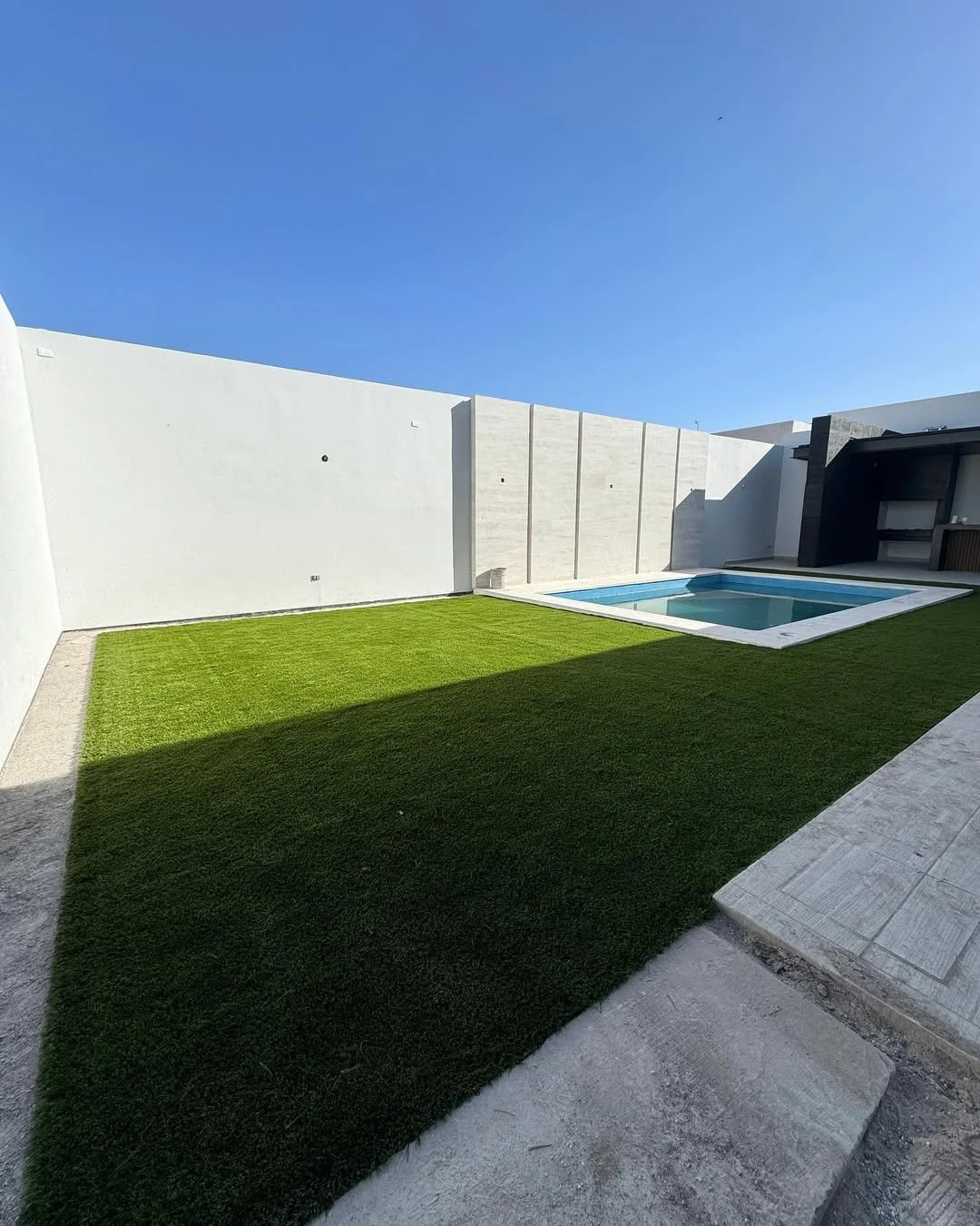 Césped artificial en un patio con una piscina pequeña y muro de cemento blanco, en un día soleado con cielo despejado.