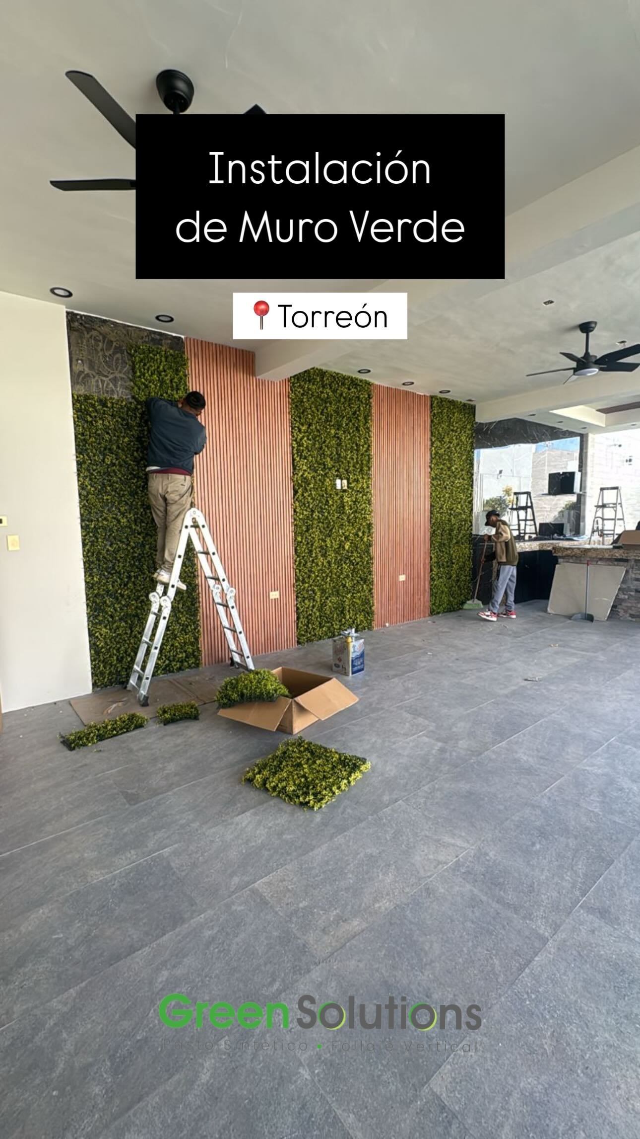 Pared decorativa con plantas verdes y listones de madera en una sala interior, durante la instalación, con dos personas trabajando y herramientas.