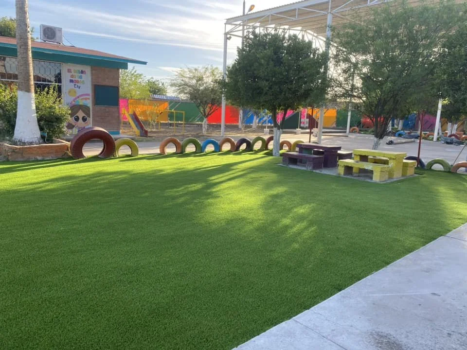 Área de juegos infantil con césped verde, árboles, un área de asientos de bloques de concreto y neumáticos decorativos, en un parque al aire libre.