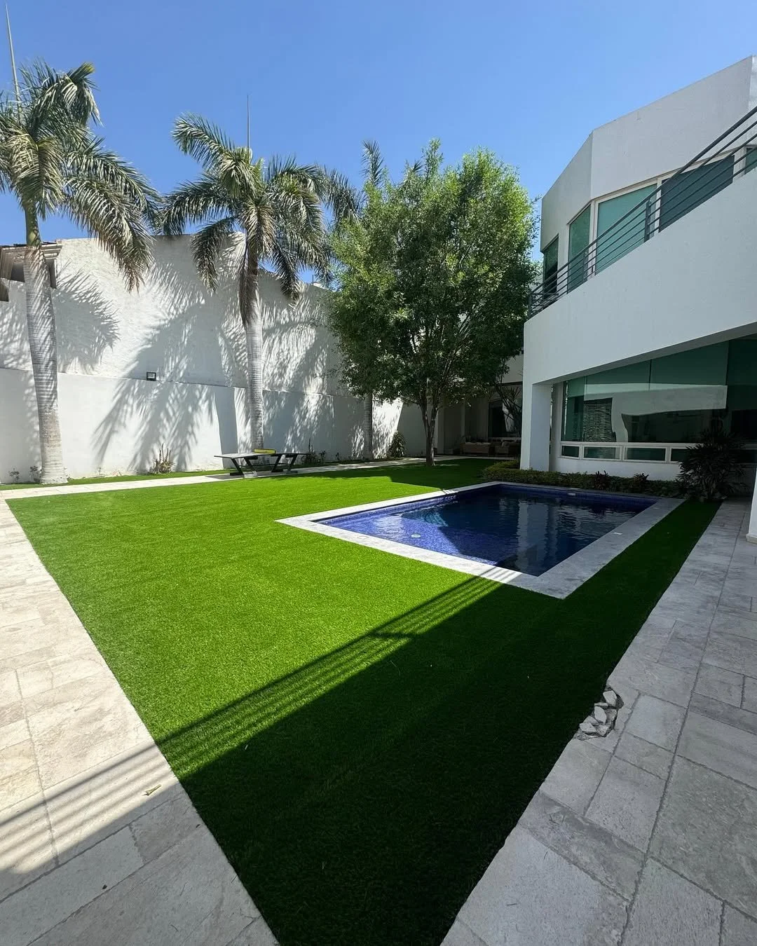 Jardín con césped verde artificial, piscina rectangular, palmeras altas, árbol frondoso, mueble de picnic y patio de piedra, casa moderna con balcón y ventanas grandes, cielo despejado.
