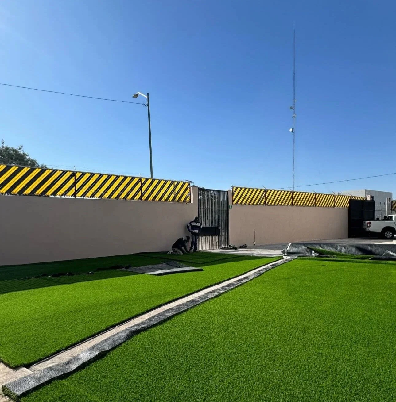 Trabajadores colocando césped artificial frente a una pared con vallas amarillas y negras en una propiedad, cielo despejado y algunas herramientas en el suelo.