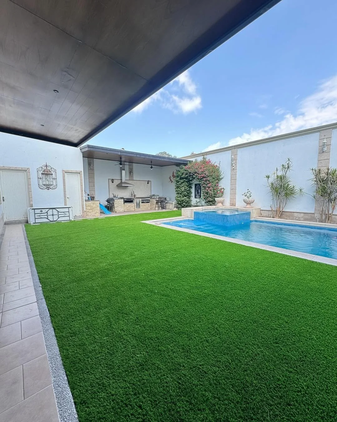 Jardín con césped artificial, piscina y área de barbacoa cubierta.