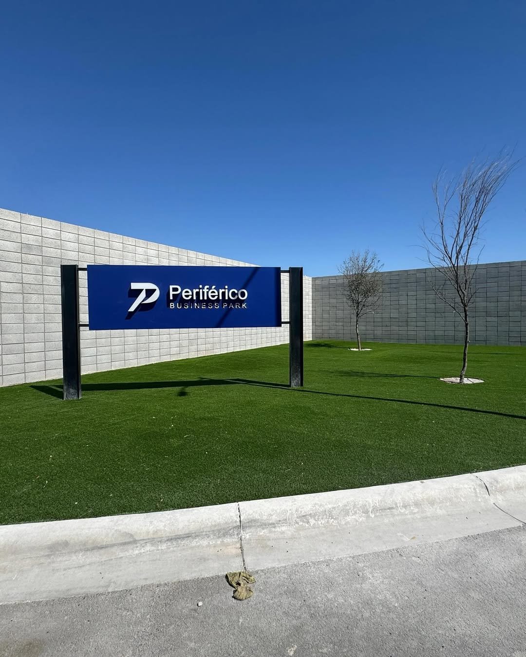 Entrada a un parque empresarial llamado 'Periférico Business Park', con un cartel azul, césped verde, pared de bloques y árboles sin hojas.