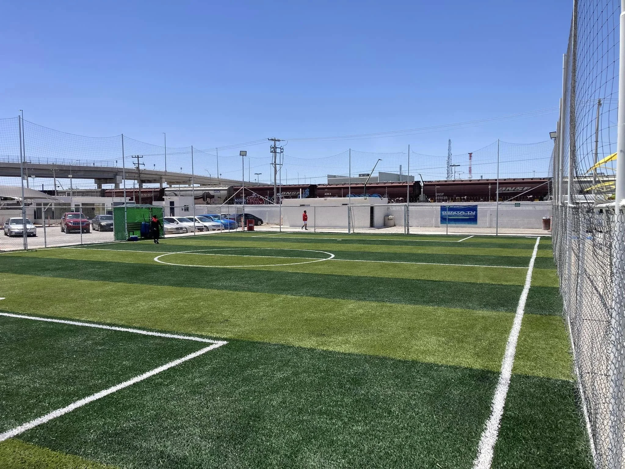 Cancha de fútbol con césped artificial en un campo cerrado con malla y fondo con autos y trenes