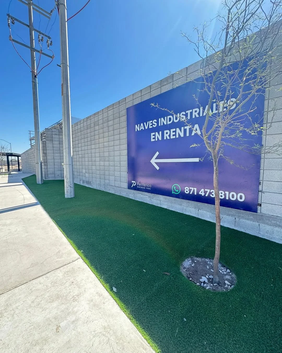 Cartel azul que dice 'Naves industriales en renta' con flecha a la izquierda, junto a un árbol y una pared de bloques de cemento, en un área con césped artificial y suelo de concreto.