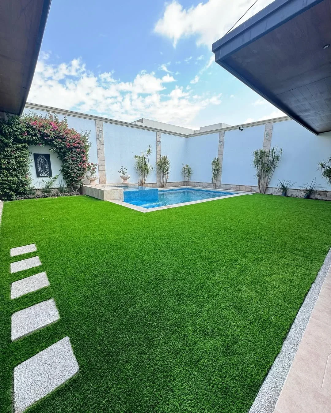Patio con césped artificial, piscina pequeña, paredes blancas, plantas y cielo con nubes.