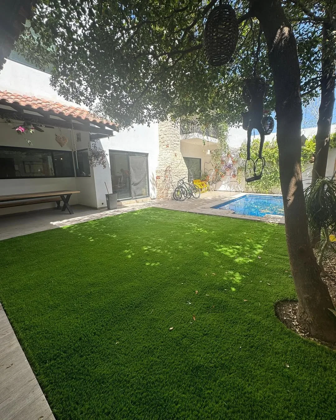 Jardín con césped artificial, árbol con columpios y colgantes, piscina pequeña, bicicletas y bancos, en un espacio exterior de casa.