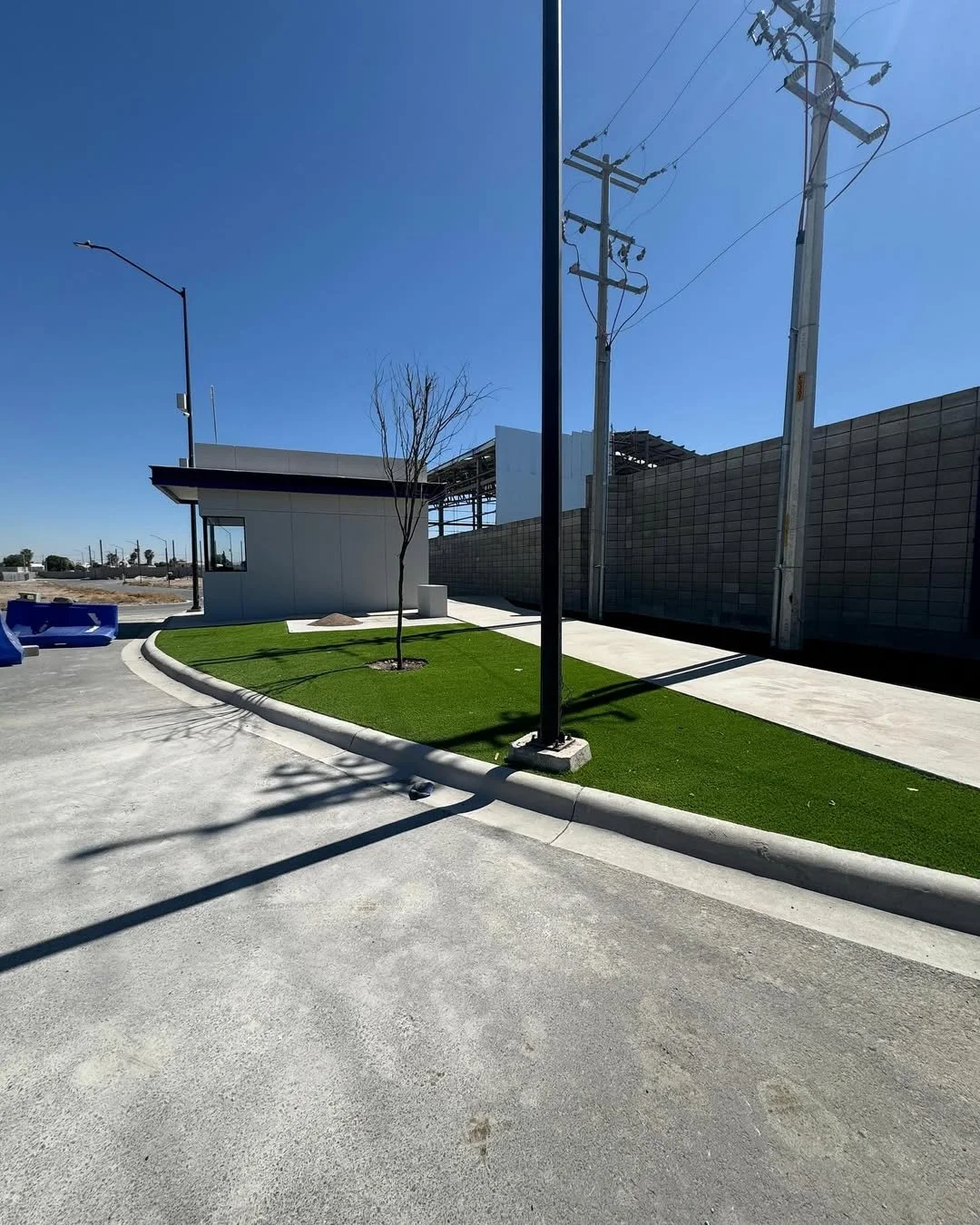 Área exterior con césped artificial, árboles, una construcción pequeña y un muro de bloques. También hay postes de electricidad y sombra en el suelo.