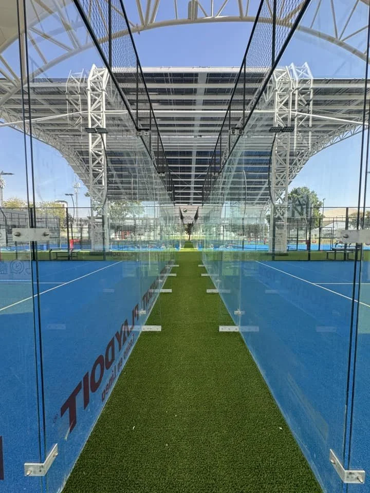 Pasillo entre canchas de pádel con estructura metálica y cristales, con césped artificial en el centro.