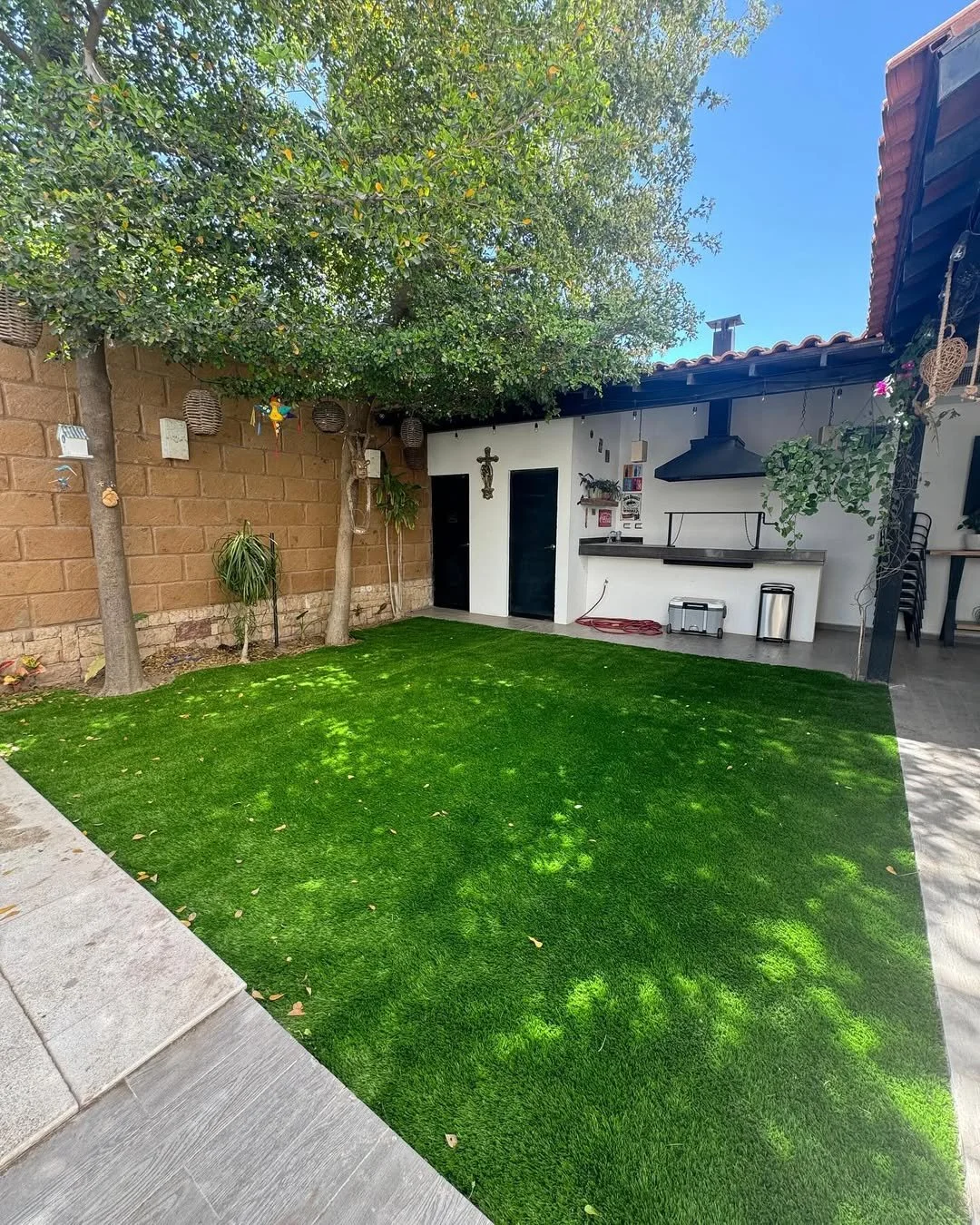 Jardín con césped artificial, árboles, pared de ladrillos, área de barbacoa blanca con sombrero negro y utensilios, y decoraciones en las paredes.