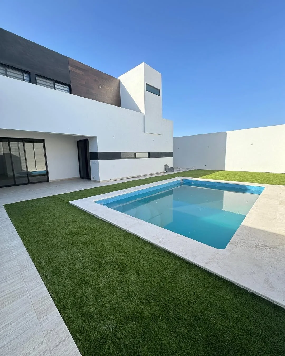 Vista de una piscina rectangular en un patio con césped artificial, paredes blancas y una casa moderna con ventanas de vidrio y paredes de color blanco y gris.