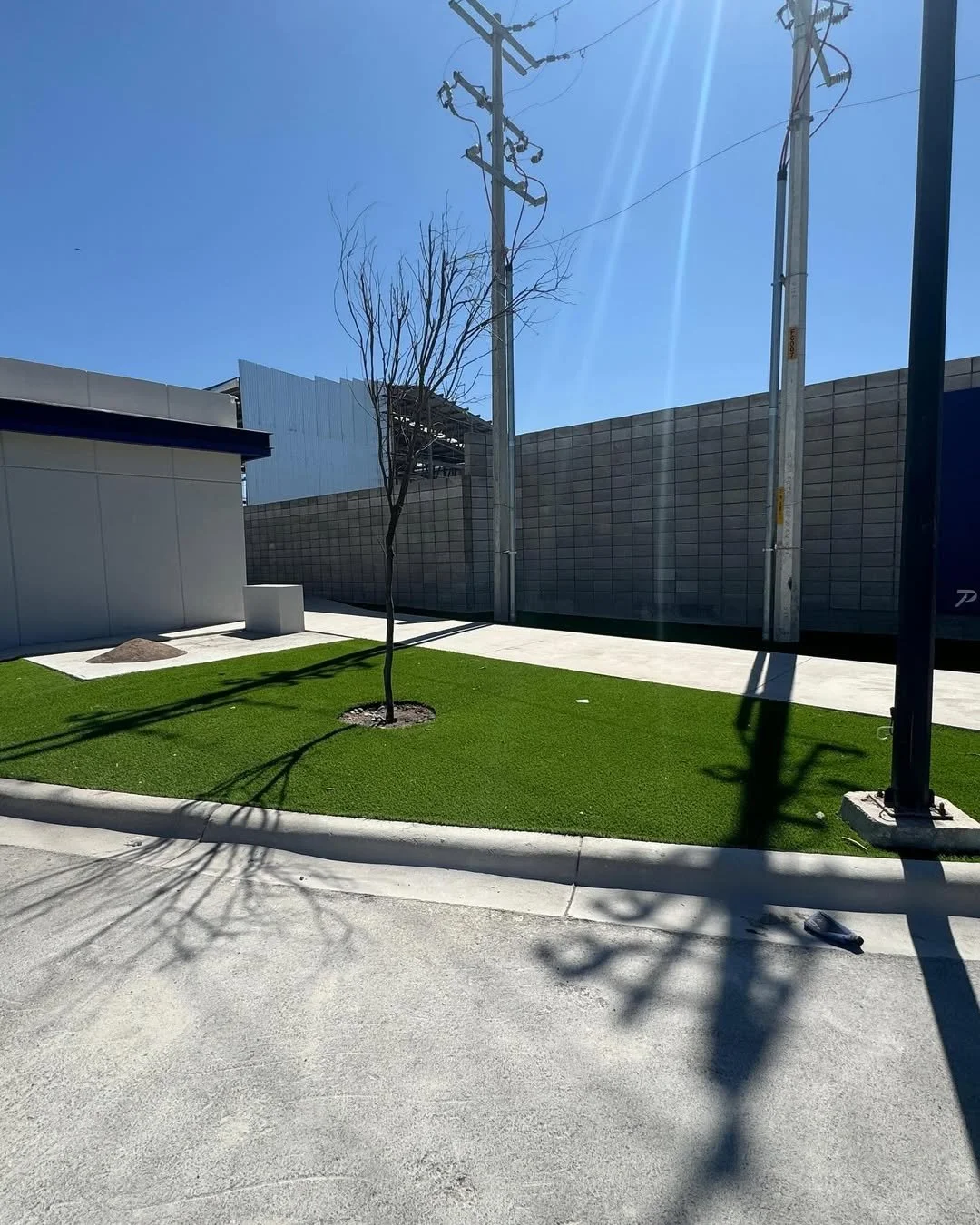 Un espacio exterior con césped artificial, un árbol sin hojas, sombra de cables y postes de electricidad, y una pared de bloques de cemento, en un día soleado.