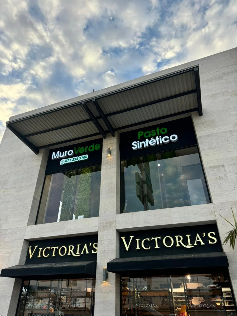 Vista de un edificio comercial con las señalizaciones de 'Muro Verde', 'Pasto Sintético' y 'Victoria's', en una noche con cielo nublado.